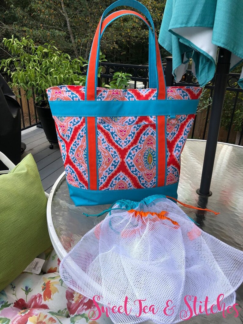 Pu&ograve; includere: Una borsa tote colorata con manici e finiture arancioni e un tessuto fantasia rosso, blu e pesca. Include due sacchetti a coulisse in rete bianca. Il testo "Sweet Tea & Stitches" &egrave; visibile.