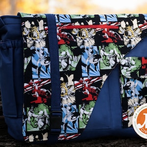 Op de afbeelding: Een blauwe canvas messenger tas met een klep sluiting. De klep is gemaakt van zwarte stof met een herhalend patroon van stripfiguren, waaronder Captain America en Hulk. De tas heeft een ritsvak aan de voorkant en een kleiner vak aan de zijkant. De tas heeft ook een band met een gesp. De tas heeft een label met de tekst "RLR Creations Sewing Patterns" en "RLR Creations - Rachel Blount Designer".