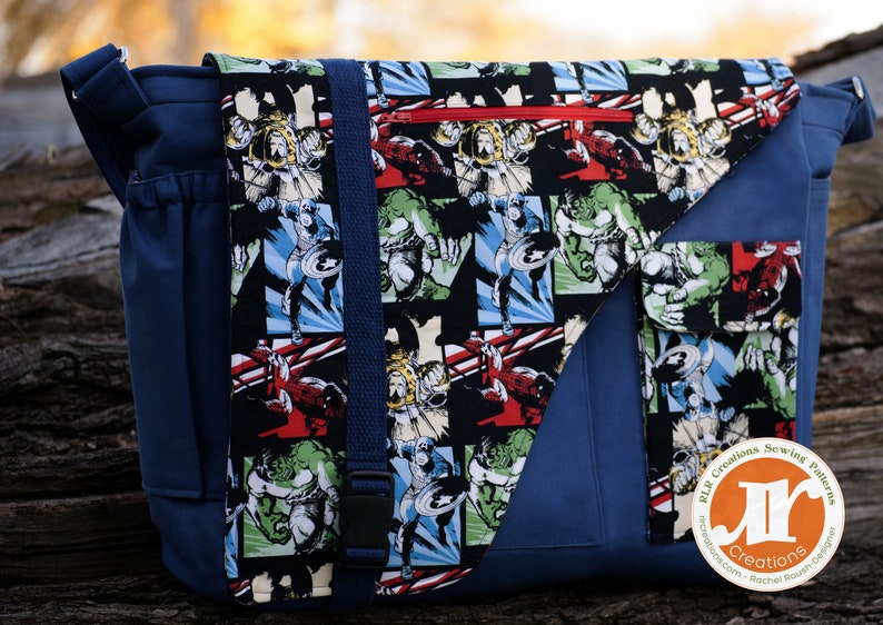 Op de afbeelding: Een blauwe canvas messenger tas met een klep sluiting. De klep is gemaakt van zwarte stof met een herhalend patroon van stripfiguren, waaronder Captain America en Hulk. De tas heeft een ritsvak aan de voorkant en een kleiner vak aan de zijkant. De tas heeft ook een band met een gesp. De tas heeft een label met de tekst "RLR Creations Sewing Patterns" en "RLR Creations - Rachel Blount Designer".