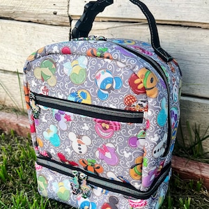 Puede incluir: Una bolsa de almuerzo gris con un asa negra y dos compartimentos con cremallera. La bolsa est&aacute; cubierta con un patr&oacute;n de personajes de Disney coloridos, incluidos Mickey Mouse, Minnie Mouse, Donald Duck y Goofy. El texto "Handcrafted by Yvonne" est&aacute; impreso en la bolsa.