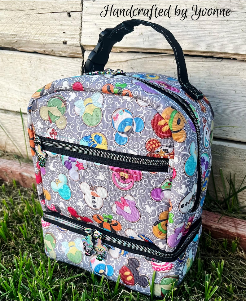 Puede incluir: Una bolsa de almuerzo gris con un asa negra y dos compartimentos con cremallera. La bolsa est&aacute; cubierta con un patr&oacute;n de personajes de Disney coloridos, incluidos Mickey Mouse, Minnie Mouse, Donald Duck y Goofy. El texto "Handcrafted by Yvonne" est&aacute; impreso en la bolsa.