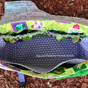 Op de afbeelding: Een groene en paarse stoffen tas met een ritssluiting bovenaan en een paarse en witte geprinte voering. De tas heeft een band en is open om de binnenkant te laten zien. De tas ligt op een bed van bruine houtsnippers. De tekst "Save water" en "Save this" is zichtbaar op de tas.