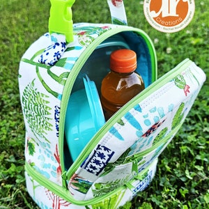 Puede incluir: Una bolsa de almuerzo verde y blanca con cierre de cremallera y asa verde. La bolsa est&aacute; abierta y contiene un recipiente azul y una botella transparente con tapa naranja. La bolsa est&aacute; sentada sobre un lecho de hierba verde.