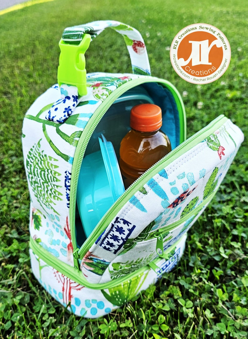 Puede incluir: Una bolsa de almuerzo verde y blanca con cierre de cremallera y asa verde. La bolsa est&aacute; abierta y contiene un recipiente azul y una botella transparente con tapa naranja. La bolsa est&aacute; sentada sobre un lecho de hierba verde.