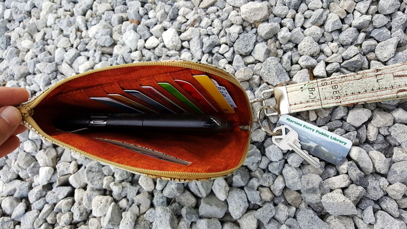 Pode incluir: Bolsa aberta com z&iacute;per com um exterior estampado em marrom e bege. O interior &eacute; forrado com tecido laranja e cont&eacute;m v&aacute;rios cart&otilde;es de cr&eacute;dito, uma caneta preta e um pequeno bolso com z&iacute;per. Um chaveiro com uma al&ccedil;a de tecido branco e marrom e um cart&atilde;o de biblioteca est&aacute; preso &agrave; bolsa.