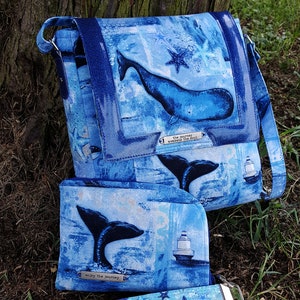 Pode incluir: Uma bolsa de m&atilde;o de tecido azul e branco com tema de baleia com uma carteira e uma al&ccedil;a combinando. A bolsa tem um design de baleia e estrela do mar com o texto "the journey awakens the soul". A carteira tem um design de cauda de baleia e farol com o texto "enjoy the journey".