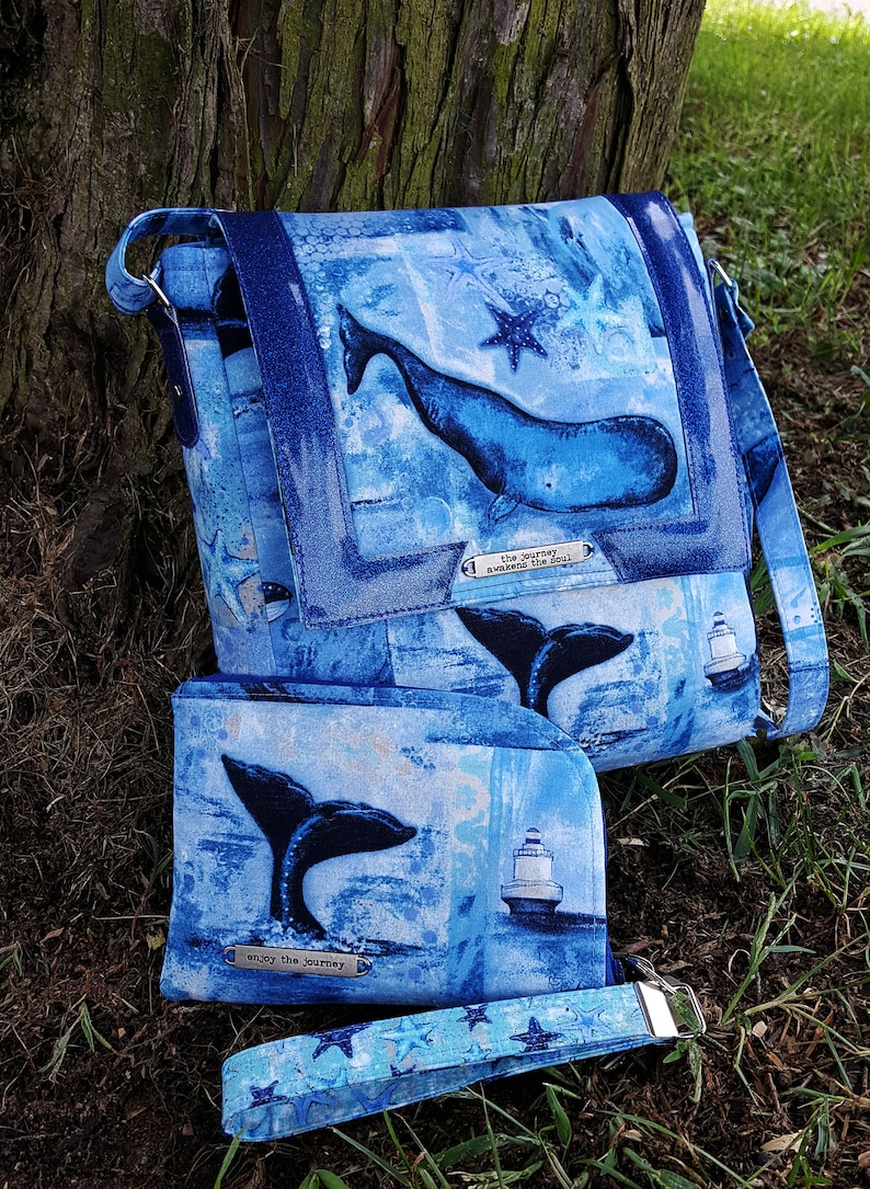 Pode incluir: Uma bolsa de m&atilde;o de tecido azul e branco com tema de baleia com uma carteira e uma al&ccedil;a combinando. A bolsa tem um design de baleia e estrela do mar com o texto "the journey awakens the soul". A carteira tem um design de cauda de baleia e farol com o texto "enjoy the journey".