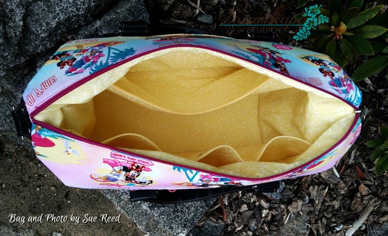 May include: A pink and blue fabric tote bag with a Mickey Mouse and Minnie Mouse surfing theme. The bag is open and shows a yellow fabric lining with multiple pockets.