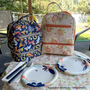 Puede incluir: Dos bolsas de almuerzo coloridas con compartimentos con cremallera. La bolsa de la izquierda tiene un fondo azul con un estampado de arco&iacute;ris y animales. La bolsa de la derecha tiene un estampado floral con detalles naranjas. Las bolsas est&aacute;n sobre una mesa con dos platos de papel y un tenedor.
