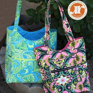 May include: Two tote bags with floral patterns. The bag on the left is green and blue with a paisley design. The bag on the right is black with pink and green flowers. Both bags have a zippered pocket on the front. The bags are sitting on a stone surface.