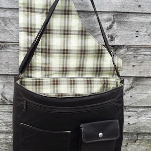 Op de afbeelding: Een zwarte canvas messenger tas met een geruite voering. De tas heeft een ritssluiting, een klep met een knoopsluiting en een groot vak aan de voorkant.