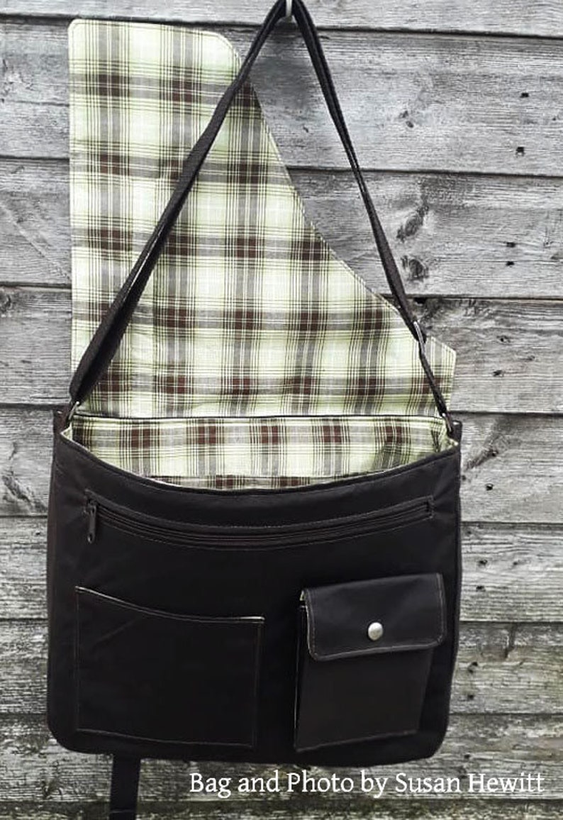 Op de afbeelding: Een zwarte canvas messenger tas met een geruite voering. De tas heeft een ritssluiting, een klep met een knoopsluiting en een groot vak aan de voorkant.