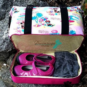 May include: A pink travel bag with a Mickey Mouse themed print featuring the text "Surf's Up!" The bag is open and shows a pair of pink shoes and a gray pair of pants inside.