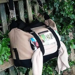 May include: A beige and black canvas tote bag with a black strap and a zippered pocket. The bag is filled with items, including a magazine and a white scarf.