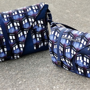 Op de afbeelding: Twee blauwe messenger bags met een wit en blauw patroon met een herhalend beeld van een politiebak. De grotere tas heeft een zwarte band en de kleinere tas heeft een zilveren rits.