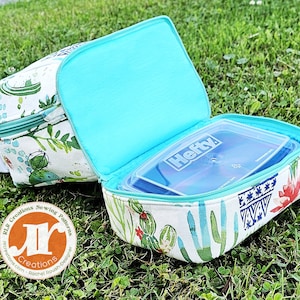Puede incluir: Una bolsa de almuerzo blanca con estampado de cactus y forro turquesa. La bolsa est&aacute; abierta y contiene un recipiente de pl&aacute;stico transparente Hefty.
