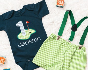 May include: A baby boy's first birthday outfit with a navy blue bodysuit featuring a golf green number one with a red flag and the name "Jackson" embroidered on it. The outfit also includes green gingham shorts and green braces.