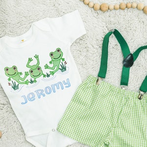 May include: A white bodysuit with a green frog design and the name "Jeromy" embroidered on it. A green gingham shorts outfit with green suspenders.