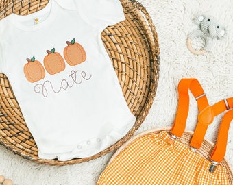 Könnte beinhalten: Ein weißer Baby-Strampler mit drei orangefarbenen Kürbissen und dem Namen "Nate", der auf der Vorderseite gestickt ist. Der Strampler liegt in einem geflochtenen Korb. Eine orangefarbene Gingham-Shorts mit orangefarbenen Hosenträgern liegt in einem geflochtenen Korb.