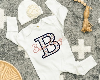 May include: White baby onesie with a floral print and a navy blue letter B with the word "Baby" in pink script.