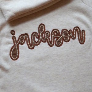 K&ouml;nnte beinhalten: Ein wei&szlig;es Shirt mit dem Namen "jackson" in braunem Garn gestickt.