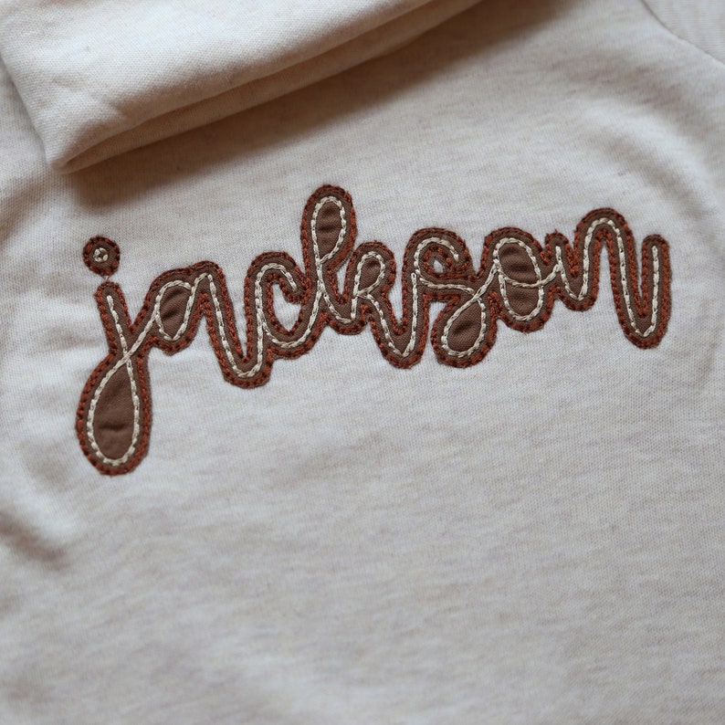 K&ouml;nnte beinhalten: Ein wei&szlig;es Shirt mit dem Namen "jackson" in braunem Garn gestickt.