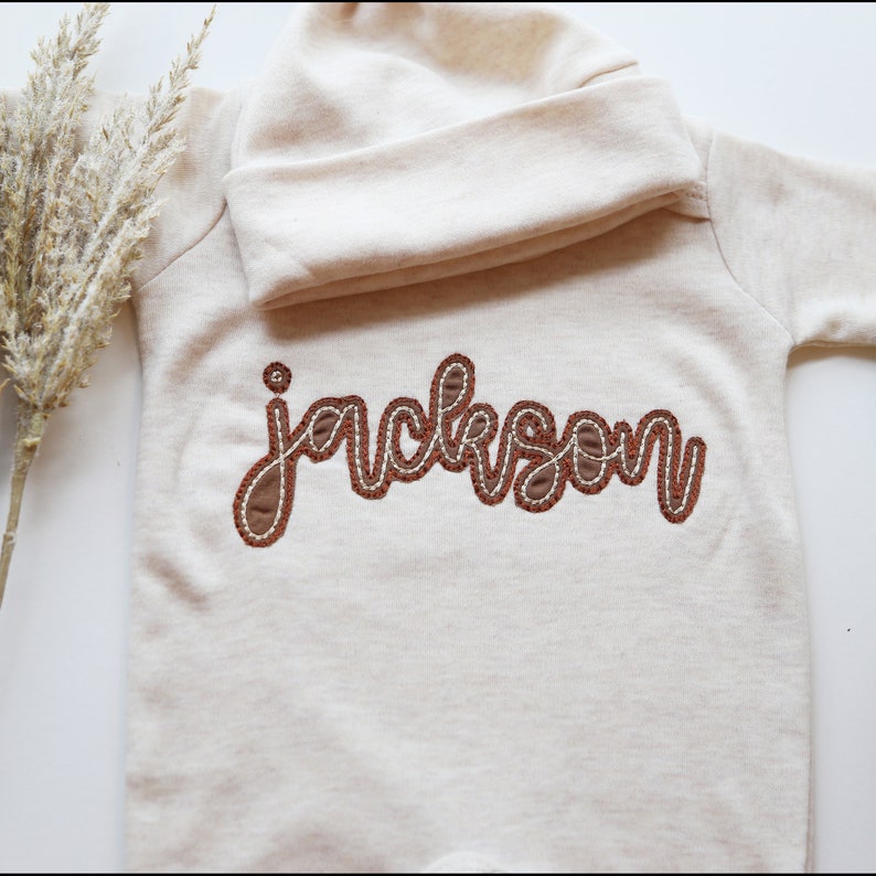 K&ouml;nnte beinhalten: Ein cremefarbener Baby-Strampler mit einem braunen gestickten Namen "Jackson" auf der Vorderseite. Der Strampler hat einen umschlagbaren Kragen.