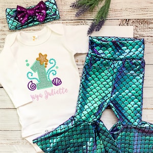 Op de afbeelding: Een baby-outfitset met zeemeerminthema. Het bevat een witte romper met een nummer 1 en een naam, een bijpassende hoofdband met een paillettenstrik en een legging met zeemeerminschaal. De set heeft teal, paarse en gouden kleuren.