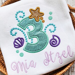 Op de afbeelding: Wit T-shirt met een zeemeerminthematisch ontwerp. Voorzien van een turquoise glitterend cijfer 3, een gouden glitter zeester en turquoise en paarse schelp- en belaccenten. De naam "Mia Itzel" is geborduurd in lavendel.