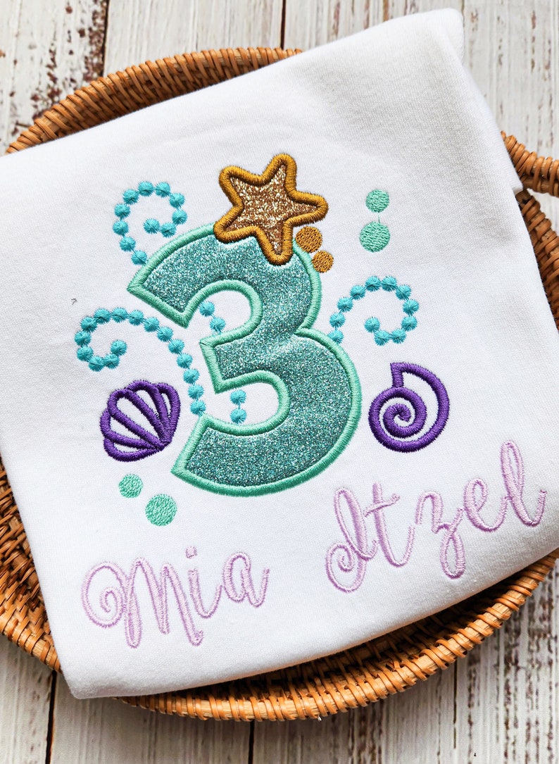 Op de afbeelding: Wit T-shirt met een zeemeerminthematisch ontwerp. Voorzien van een turquoise glitterend cijfer 3, een gouden glitter zeester en turquoise en paarse schelp- en belaccenten. De naam "Mia Itzel" is geborduurd in lavendel.