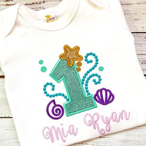 Op de afbeelding: Witte babybody met een turquoise glitterende cijfer 1, een goudkleurige glitterster en een paars schelpmotief. De naam "Mia Ryan" is geborduurd in lichtpaars schrift. Voor een eerste verjaardag.