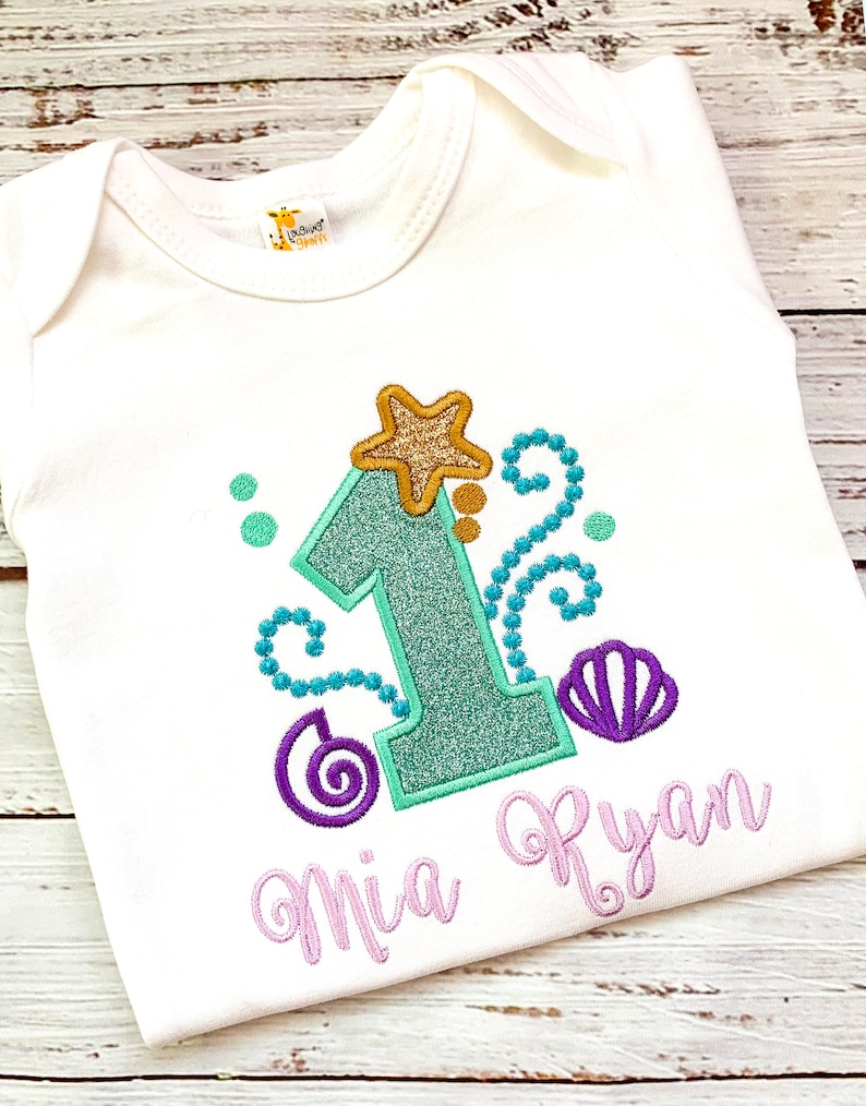 Op de afbeelding: Witte babybody met een turquoise glitterende cijfer 1, een goudkleurige glitterster en een paars schelpmotief. De naam "Mia Ryan" is geborduurd in lichtpaars schrift. Voor een eerste verjaardag.