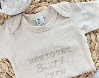 May include: A beige baby onesie with the text "New to the Smith Crew" embroidered in gold thread.