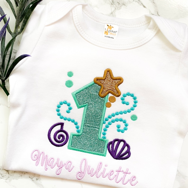 Op de afbeelding: Witte babybody met een turquoise glitter nummer &eacute;&eacute;n, een goudkleurige glitter zeester en turquoise en paarse schelpaccenten. De naam "Maya Juliette" is geborduurd in lavendelkleurig schrift. Een verjaardagsoutfit.