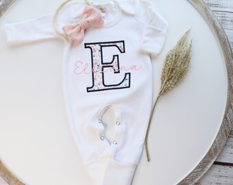 May include: White baby onesie with a floral print letter E on the front. The onesie has long sleeves and snaps at the crotch. A pink bow headband is included.