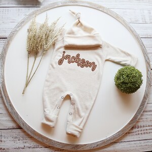 K&ouml;nnte beinhalten: Ein beiger Baby-Strampler mit langen &Auml;rmeln und einer passenden M&uuml;tze. Der Strampler hat den Namen "Jackson" in braunem Garn gestickt.