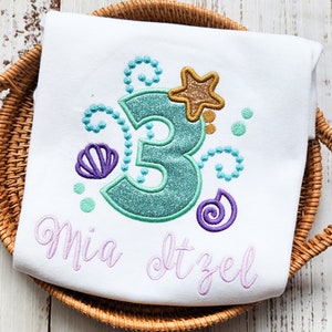 Puede incluir: Camiseta blanca con un n&uacute;mero 3 turquesa, una concha de mar morada, una estrella dorada y remolinos morados. El texto "Mia dtzel" est&aacute; bordado en morado debajo del n&uacute;mero.