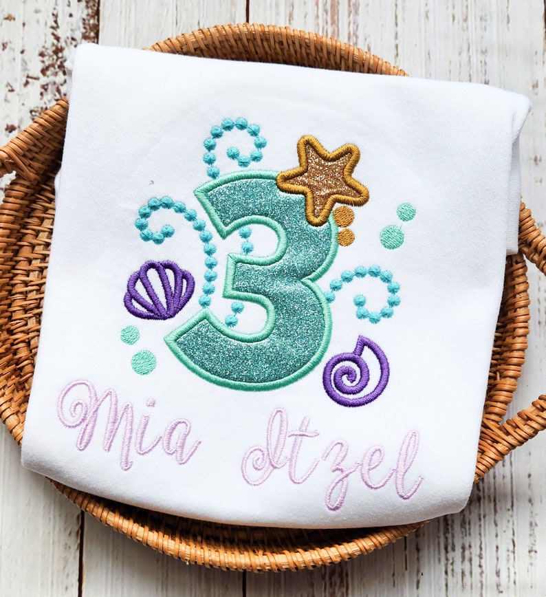 Puede incluir: Camiseta blanca con un n&uacute;mero 3 turquesa, una concha de mar morada, una estrella dorada y remolinos morados. El texto "Mia dtzel" est&aacute; bordado en morado debajo del n&uacute;mero.