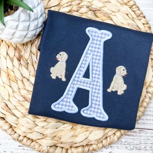 May include: A navy blue shirt with a blue and white gingham letter A and two embroidered brown dogs.