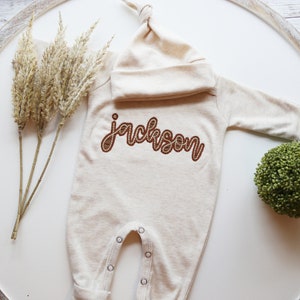 K&ouml;nnte beinhalten: Ein beiger Baby-Strampler mit langen &Auml;rmeln und einer passenden M&uuml;tze. Der Strampler hat den Namen "Jackson" in braunem Garn gestickt.