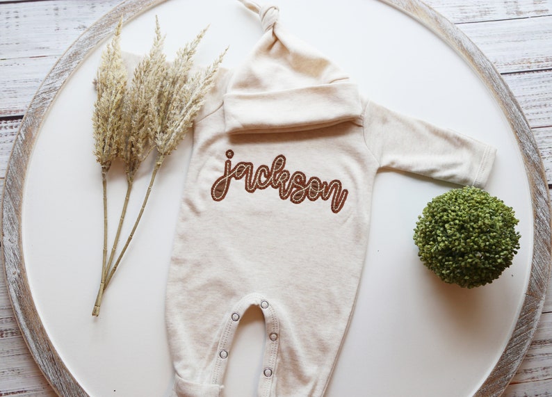 K&ouml;nnte beinhalten: Ein beiger Baby-Strampler mit langen &Auml;rmeln und einer passenden M&uuml;tze. Der Strampler hat den Namen "Jackson" in braunem Garn gestickt.