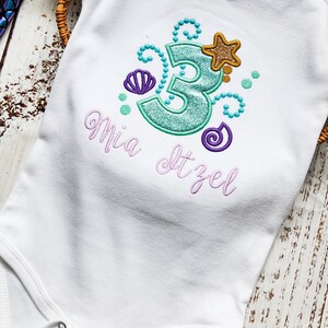 Puede incluir: Un body blanco para beb&eacute; con un n&uacute;mero 3 turquesa, una concha de mar morada, una estrella dorada y remolinos morados. El body tiene el nombre "Mia" y el texto "Itzel" bordado.