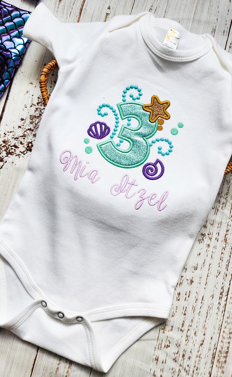 Puede incluir: Un body blanco para beb&eacute; con un n&uacute;mero 3 turquesa, una concha de mar morada, una estrella dorada y remolinos morados. El body tiene el nombre "Mia" y el texto "Itzel" bordado.