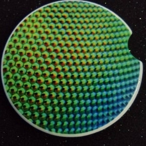 May include: A round, white coaster with a green, blue, and red iridescent pattern. The pattern resembles a honeycomb or fish scales.
