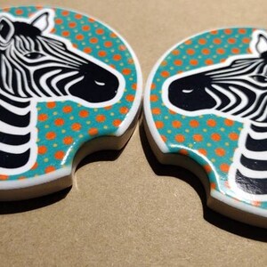 May include: Two round ceramic coasters with a teal background and orange polka dots. Each coaster features a black and white zebra head graphic.