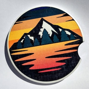 May include: A round coaster with a white background and a black and white grid pattern. The design features a mountain range with a snowy peak, surrounded by trees and a reflection in a lake. The colors are orange, yellow, blue, and green.