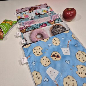 May include: A selection of reusable snack bags with diverse patterns, including cookies, cupcakes, and donuts. A red apple and a snack package are also visible. The bags have zip closures and small white tags. Designed for food storage.
