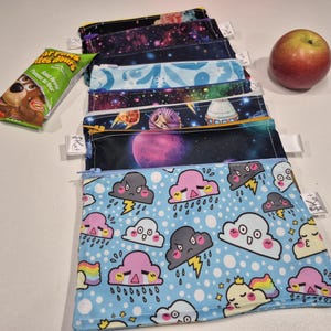 May include: A collection of reusable snack bags featuring space and cloud designs. The bags have zip closures and white tags. A red apple and a snack packet are also visible.