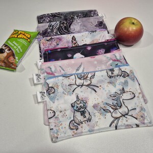 May include: A collection of reusable snack bags with diverse patterns, including unicorns and abstract designs. A green snack bar and a red apple are also visible. The bags have white tags attached. The bags are designed for carrying snacks.