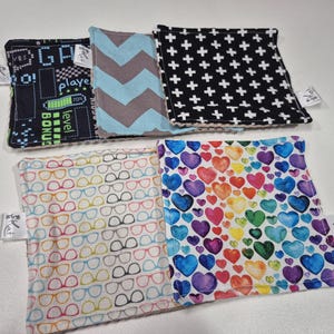 Peut inclure: Plusieurs carrés de tissu colorés avec différents motifs. On retrouve un fond noir avec des croix blanches, un motif chevron bleu et gris, un motif de cœurs arc-en-ciel et un motif de lunettes. Chaque carré a une petite étiquette.